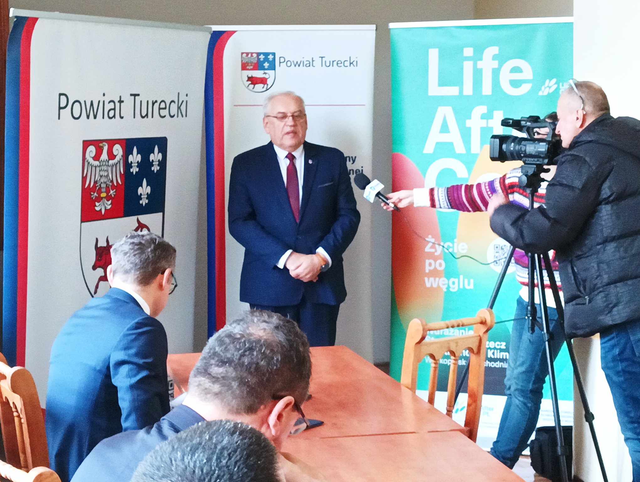 Jedna osoba stoi i mówi do kamery na tle bannerów reklamowych.