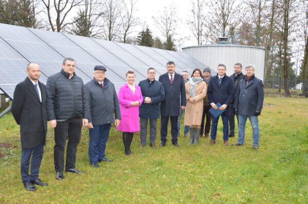 12 osób stoi na tle instalacji fotowoltaicznej, która była oficjalnie otwierana.