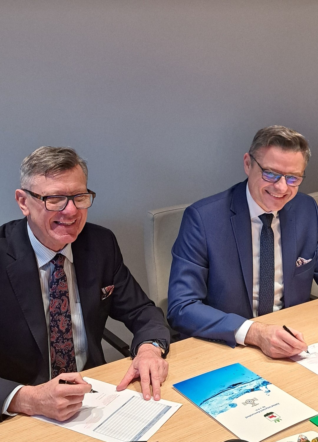 Dwie osoby siedzą na krzesłach przy stole i podpisują dokumenty.