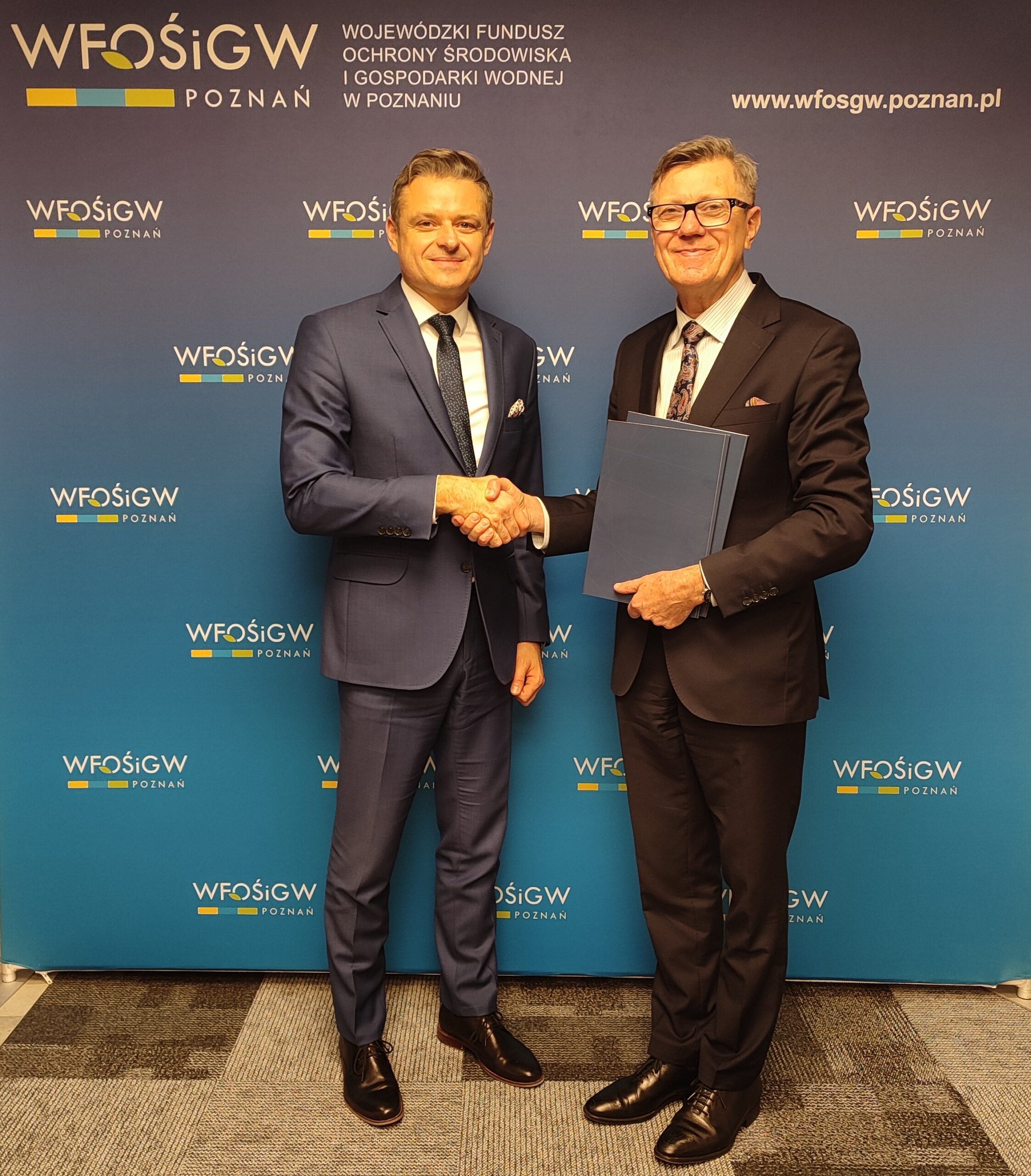 Dwie osoby stoją na tle ścianki reklamowej i podają sobie dłonie.