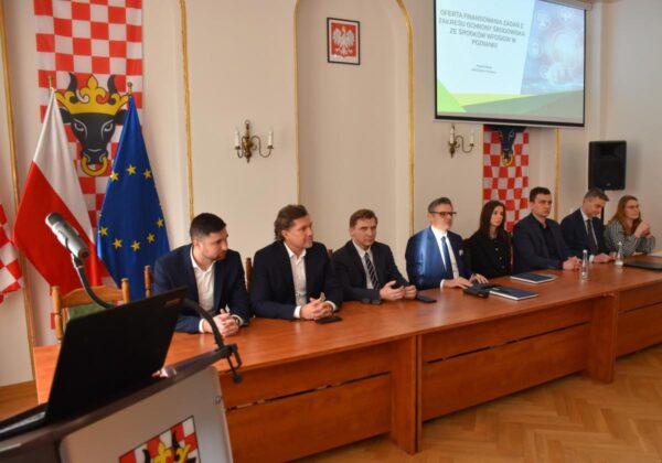 Zdjęcie przedstawia salę w urzędzie, na której za dużym stołem siedzą osoby i rozmawiają.