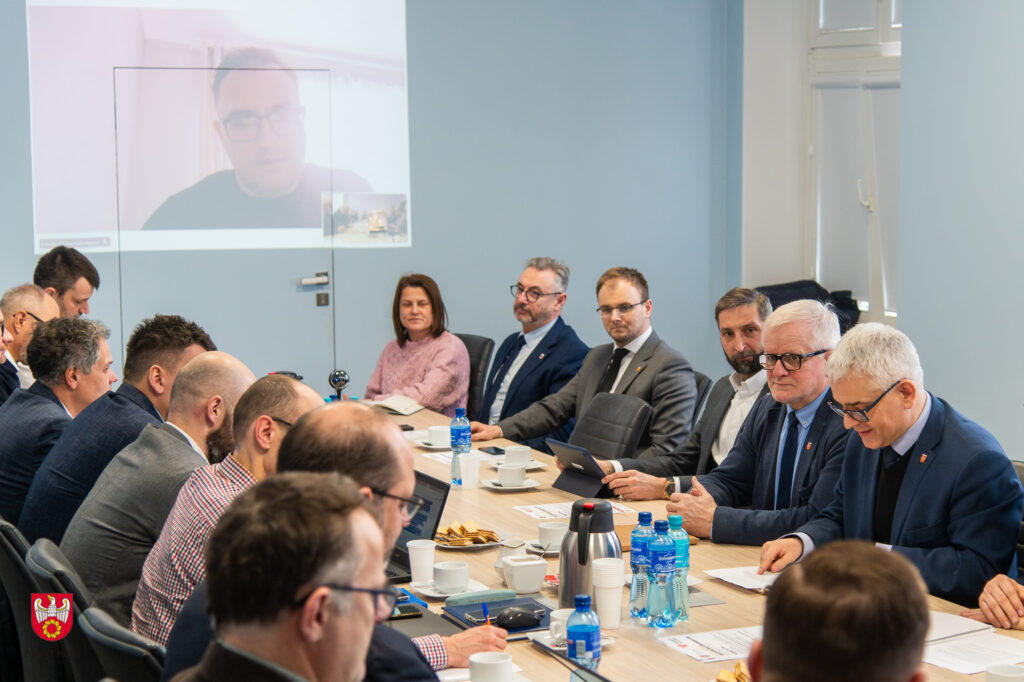 Na zdjęciu przy długim stole siedzą naprzeciwko siebie osoby biorące udział w spotkaniu.