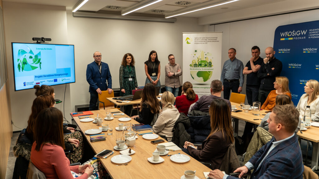 Na zdjęciu widzimy osoby zgromadzone w salce konferencyjnej, siedzące za długim stołem oraz osoby wygłaszające prelekcję, na ekranie wyświetlona jest prezentacja.