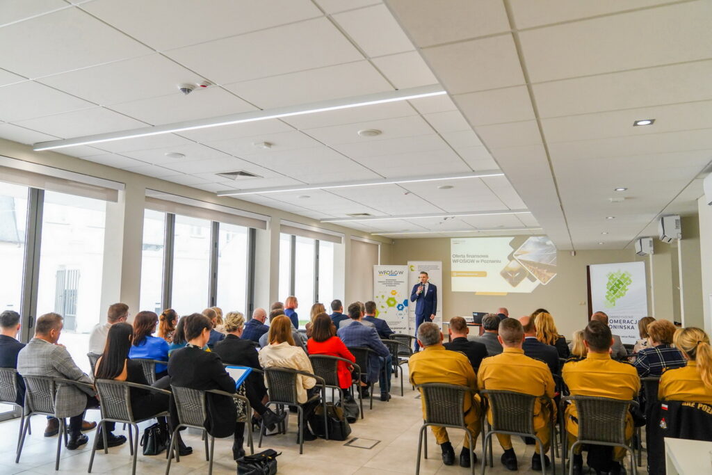 Widok na salę, w której odbywa się spotkanie - widać ludzi siedzących na krzesłąch ustawionych w rzędach, w dwóch kolumnach, a na przodzie sali widać wyświetloną prezentacje z ofertą finansową Funduszu.