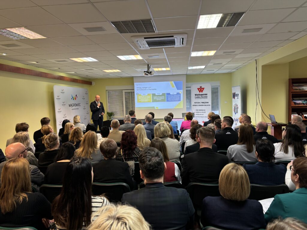 Na zdjęciu sala wypełniona ludźmi, którzy siedzą na krzesłach, a na przodzie sali stoi jedna osoba na tle banerów reklamowych i omawia przedstawioną na ekranie prezentację.