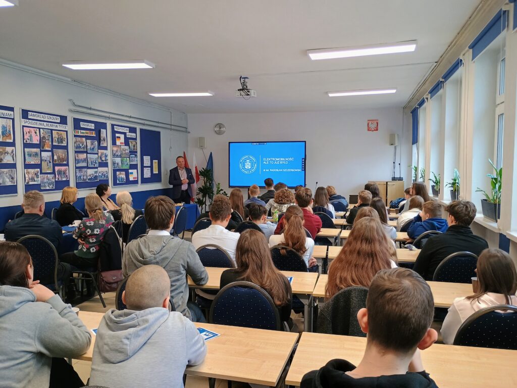 Na zdjęciu widok sali, w której odbywa się finał konkursu. Uczniowie siedzą w ławkach i słuchają wykładu. Na środku sali stoi prowadzący, a na ekranie wyświetlana jest prezentacja.