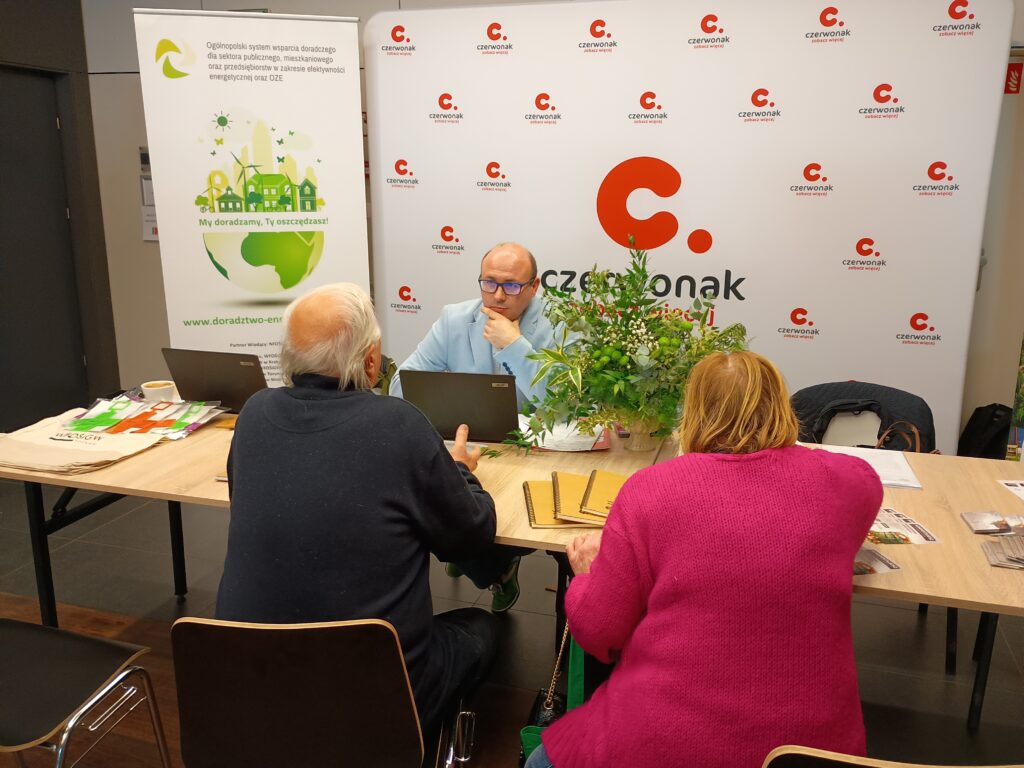 Na stoisku dwie osoby siedzą przy biurku, a jedna osoba za biurkiem i rozmawiają.