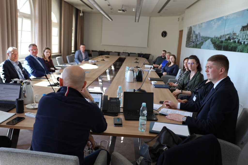 Zdjęcie przedstawia salę, a na niej wokół stołu siedzą osoby i rozmawiają.