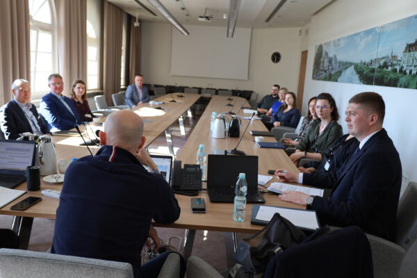 Zdjęcie przedstawia salę, a na niej wokół stołu siedzą osoby i rozmawiają.