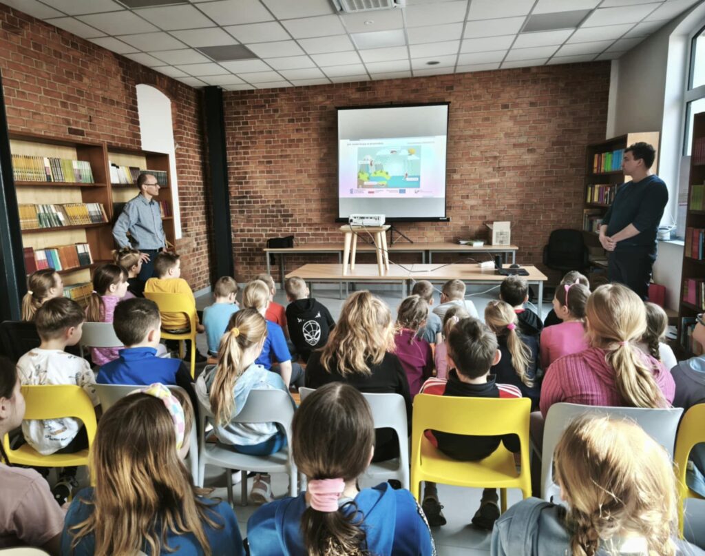 Zdjęcie przedstawia salę, w której siedzą uczniowie, a na przodzie sali widać ekran z wyświetloną prezentacją.