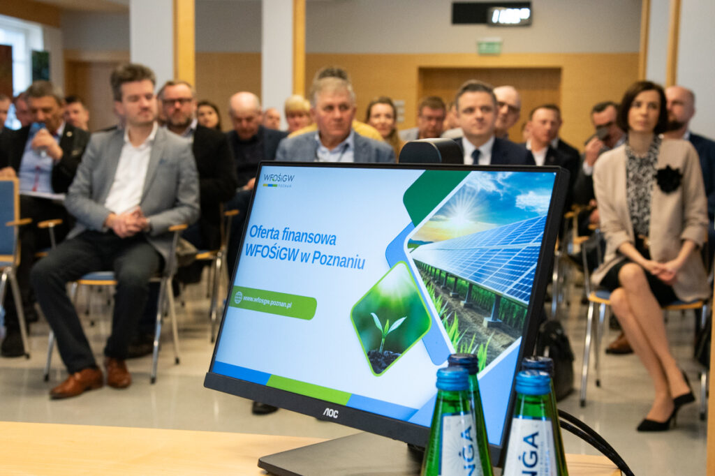 Zdjęcie przedstawia widok ekranu, na którym wyświetlona jest prezentacja, a w tle widzimy widok sali, na której na krzesłach siedzą przedstawiciele samorządów.