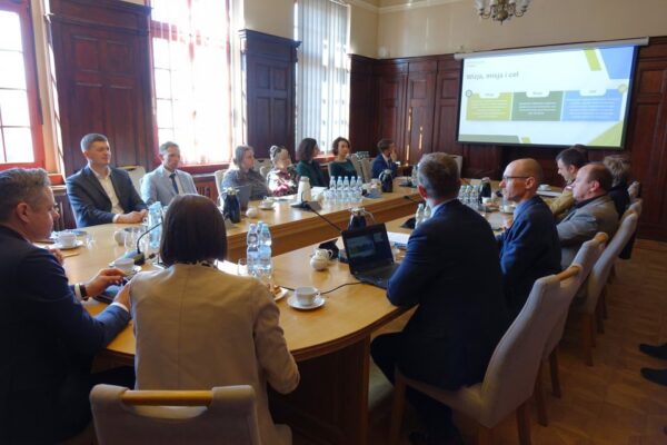 Na zdjęciu widać owalny stół, wokół którego siedzą uczesnicy spotkania, w tle widać duży ekran z wyświetloną prezentacją.