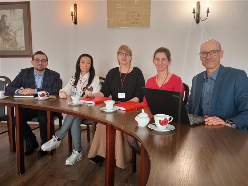 Na zdjęciu przy zaokrąglonym stole siedzi 5 osób przed laptopem i omawiają projekty.