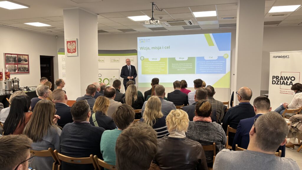 Zdjęcie przedstawia salę wypełnioną ludźmi siedzącymi na krzesłach, na środku sali ekran z wyświetloną prezentacją oraz bannery reklamowe, a przed nimi stoi jedna osoba i omawia prezentację.