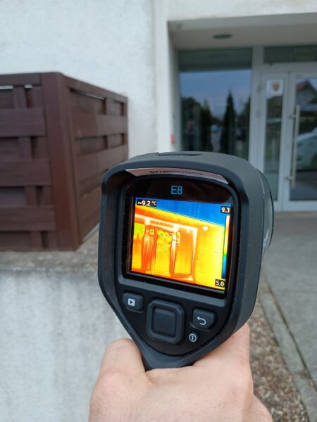 Kamera termowizyjna trzymana w dłoni na tle budynku mieszkalnego.