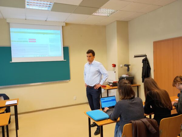 Jedna osoba stoi na środku sali wykładowej, w ławkach siedzą studenci, a przed nimi laptopy.