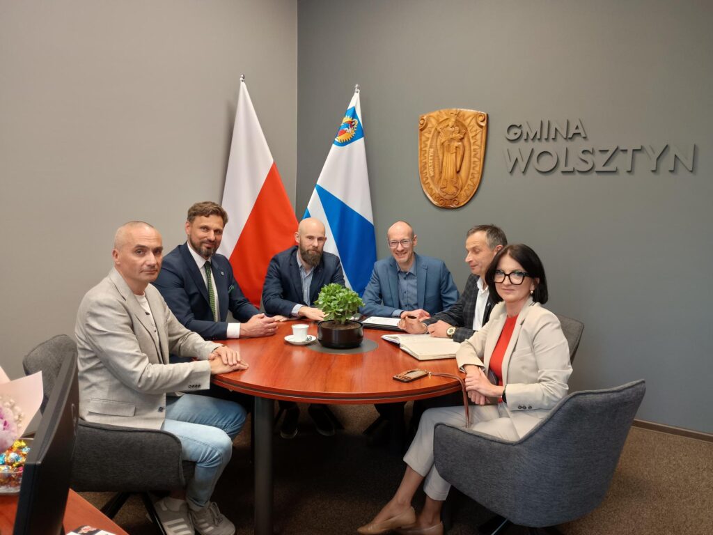 ^ osób siedzi wokół stołu, w tle widać flagę Polski oraz flagę Gminy Wolsztyn.