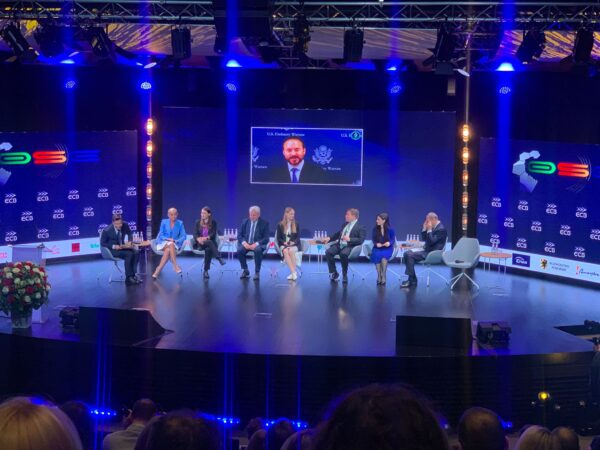 Sala, w której odbywa się wydarzenie i widok na scenę, na której siedzą na krzesłach ustawionych w stronę widowni, uczestnicy debaty. Łącznie siedzi 8 osób, na górze za nimi widać ekran, na którym widać osobę, która łączy się zdalnie z salą.