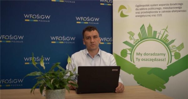 Osoba prowadząca webinar siedzi za stołem z laptopem na tle bannerów reklamowych i mówi do kamery.