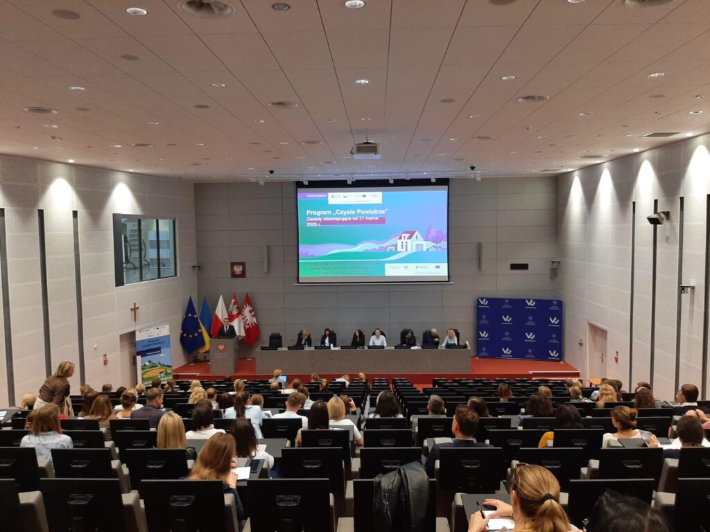 Zdjęcie przedstawia salę, na której odbywa się szkolenie. Widać od tyłu całą salę, na której siedzą uczestnicy szkolenia, i scenę, na której jedna osoba stoi za mównicą i omawia wyświetlaną prezentację.