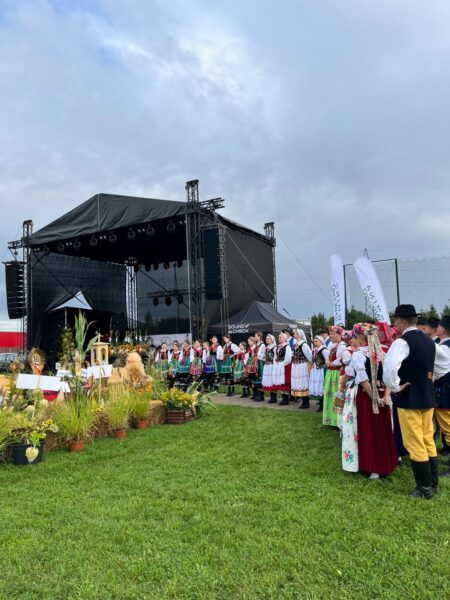 Scena podczas dożynek, a przed nią ustawiony korowód oraz przyniesione ozdoby ze słomy przygotowane specjalnie na dożynki.