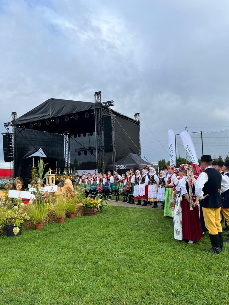 Scena podczas dożynek, a przed nią ustawiony korowód oraz przyniesione ozdoby ze słomy przygotowane specjalnie na dożynki.