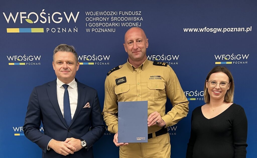 Zdjęcie przedstawia 3 osoby , które stoją na tle ścianki reklamowej z teczką z podpisaną umową o dofinansowanie.