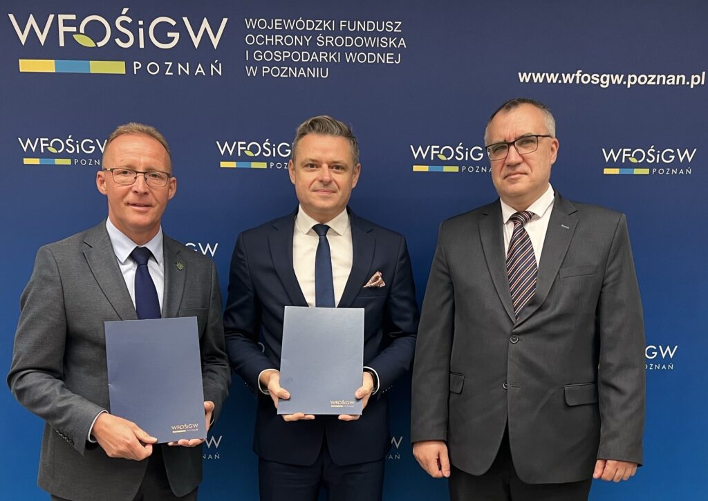 Zdjęcie przedstawia 3 osoby , które stoją na tle ścianki reklamowej z teczką z podpisaną umową o dofinansowanie.