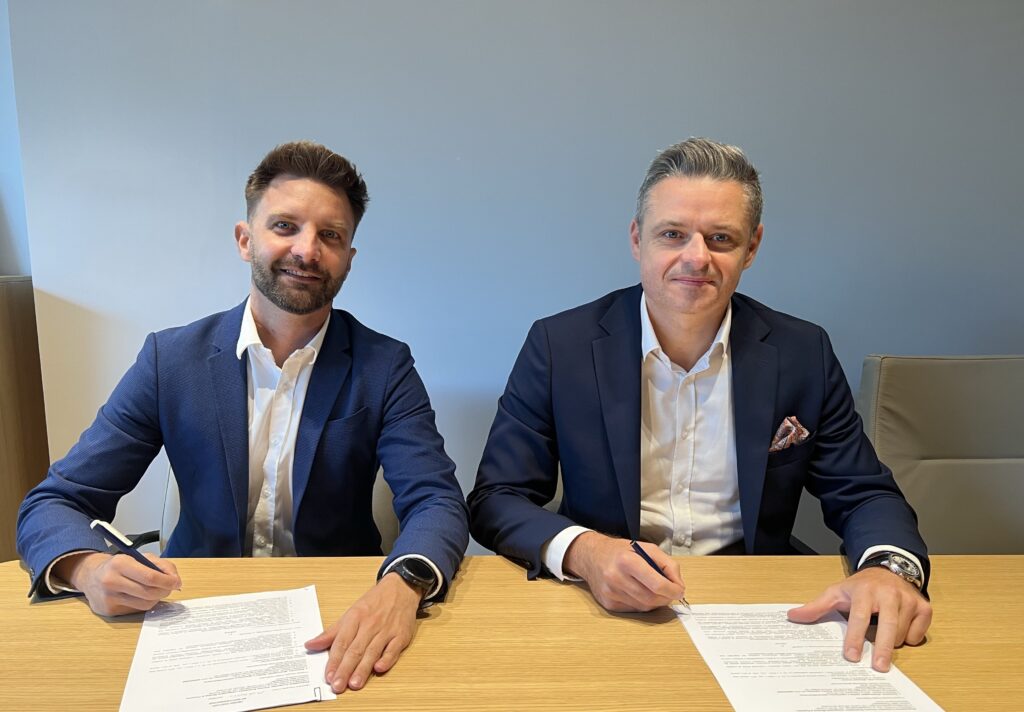 Na zdjęciu dwie osoby siedzą za stołem i podpisują umowę.