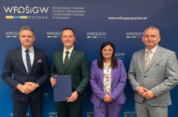 Na zdjęciu cztery osoby stoją na tle ścianki reklamowej i pozują, jedna z nich trzyma w ręku teczkę z umową.