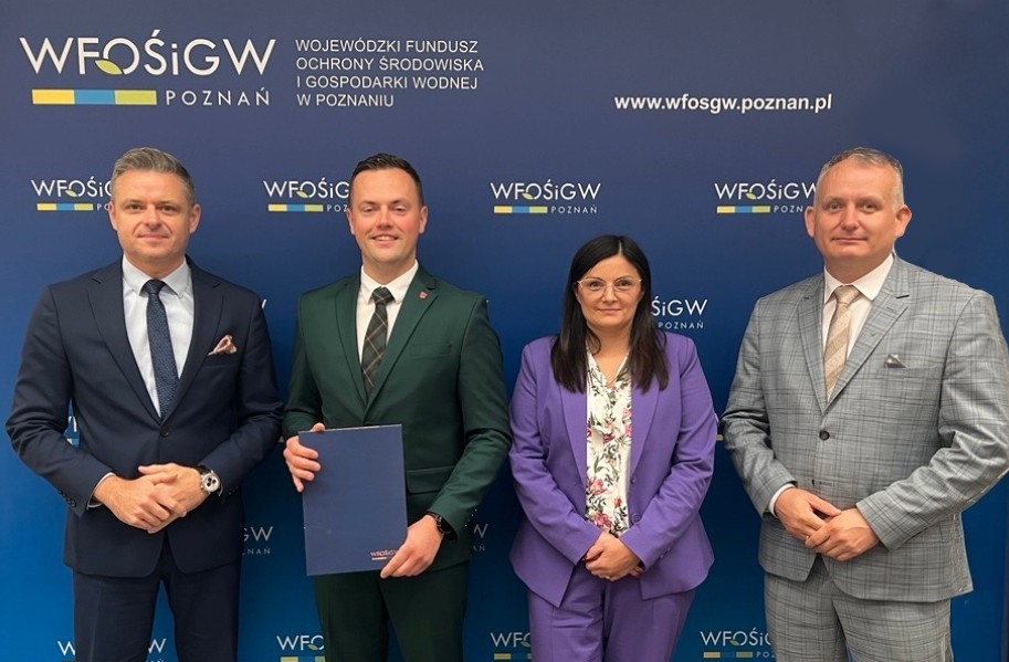 Na zdjęciu cztery osoby stoją na tle ścianki reklamowej i pozują, jedna z nich trzyma w ręku teczkę z umową.