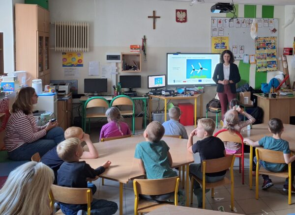Zdjęcie przedstawia klasę szkolną, w ławkach siedzą dzieci, a na środku klasy przy tablicy stoi apani i opowiada dzieciom.