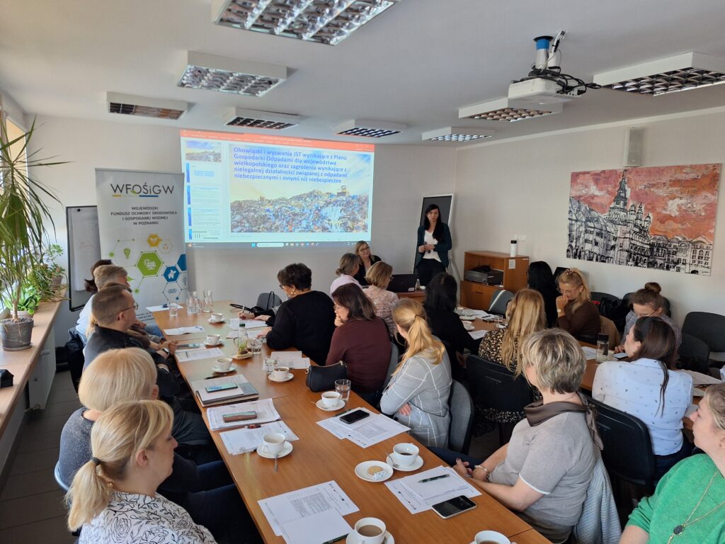 Grupa osób siedzi za stołami podczas spotkania i słucha omawianej prezentacji.