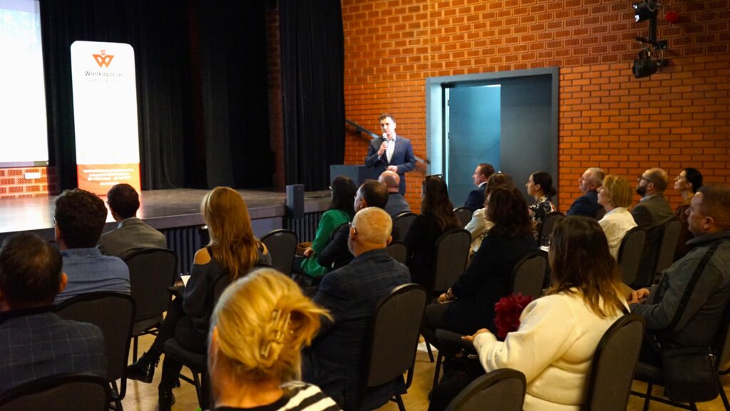 Jedna osoba stoi przed sceną i przemawia przez mikrofon do osób siedzących na sali.