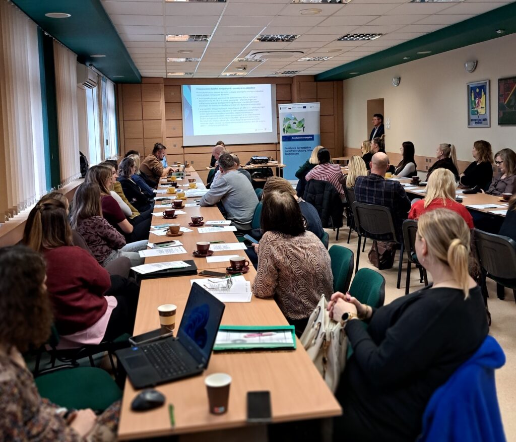 przy dwóch długich stołach siedzą uczestnicywydarzenia, a na przodziesali wyświetlana jest prezentacja i stoi osoba, która omawia prezentację.