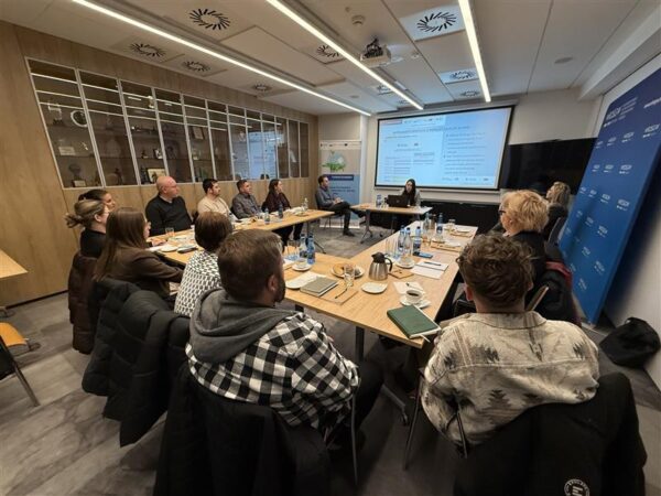 Zdjęcie przedstawia salę, na której odbywa się szkolenie. Wokół stołu ustawionego w podkowę siedzą uczestnicy szkolenia. Na początku sali za biurkiem siedzi prowadzący szkolenie, a za nim na ekranie projektora wyświetlana jest prezentacja szkoleniowa.