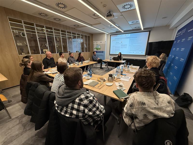 Zdjęcie przedstawia salę, na której odbywa się szkolenie. Wokół stołu ustawionego w podkowę siedzą uczestnicy szkolenia. Na początku sali za biurkiem siedzi prowadzący szkolenie, a za nim na ekranie projektora wyświetlana jest prezentacja szkoleniowa.