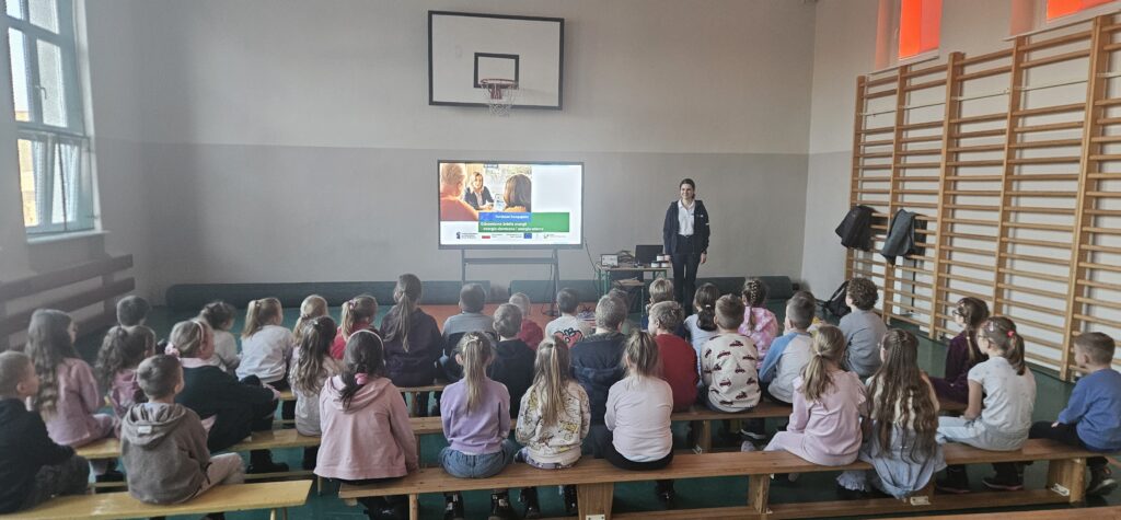 Na zdjęciu uczniowie w szkole podstawowej siedzą na ławkach na sali gimnastycznej i słuchają prelekcji. Na początku sali stoi duży telewizor, na którym wyświetlana jest prezentacja.