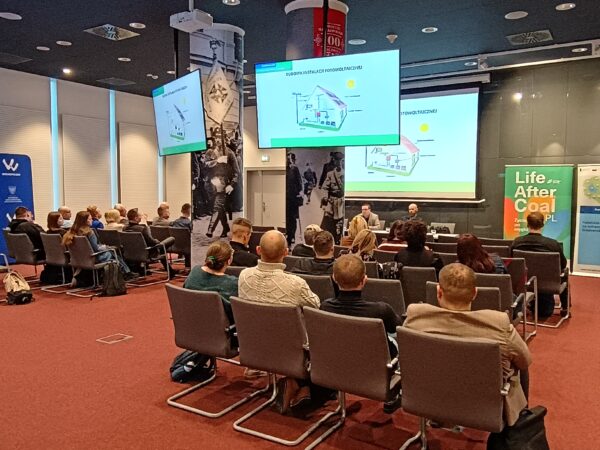 Na sali w ustawieniu kinowym siedzą uczestnicy szkolenia, a na środku sali przed ekranem, na którym wyświetlana jest prezentacja szkoleniowa stoi osoba prowadząca szkolenie.