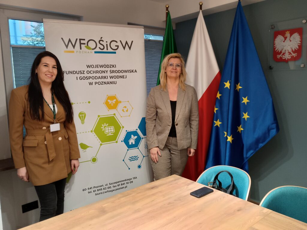 2 osoby stoją w sali przed stołem na tle flag oraz bannera reklamowego.