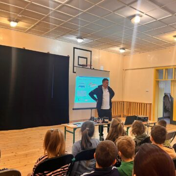 Dzieci siedzą na dużej sali gimnastycznej na krzesłach, a przed nimi stoi ekran, na którym wyświetlana jest prezentacja. Obok stoi osoba prowadząca zajęcia - doradca klimatyczno-energetyczny z WFOŚiGW w Poznaniu.