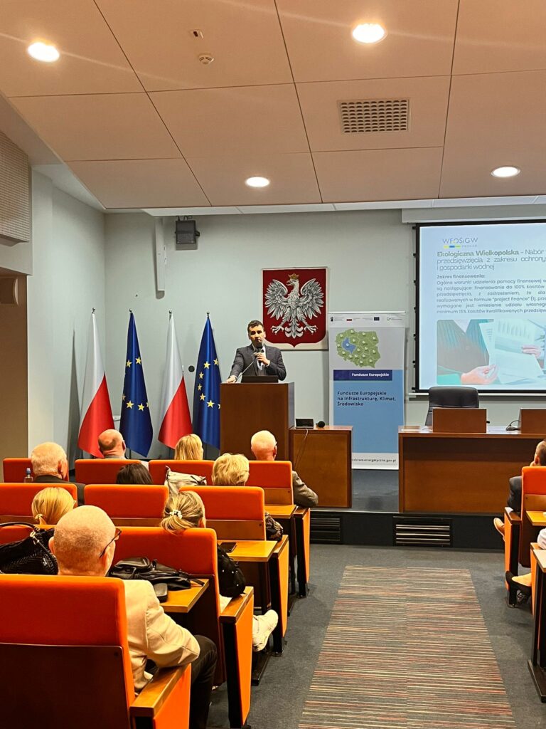 Jedna osoba stoi przy mównicy i przemawia do gości zgromadzonych na sali. Z tyłu widać banner reklamowy oraz ekran z wyświetloną prezentacją.