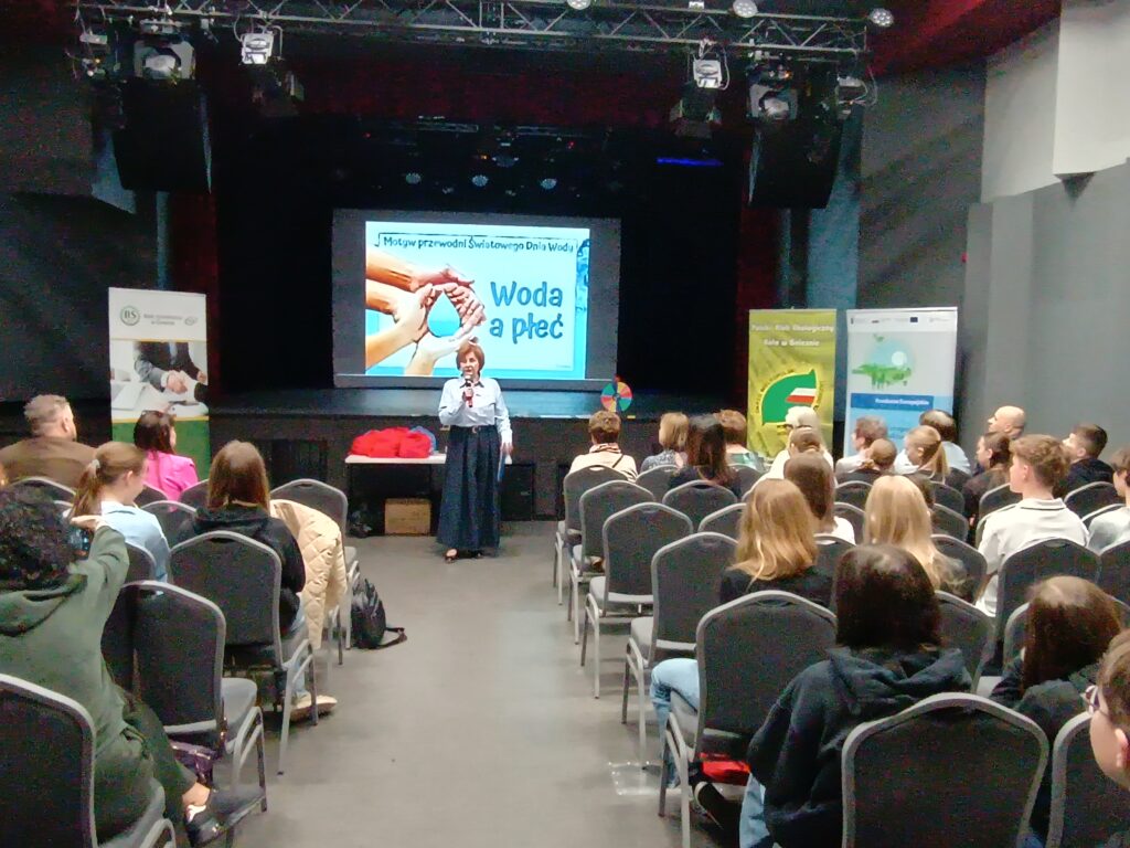 Sala wypełniona ludźmi siedzącymi na krzesłach w ustawieniu kinowym. Na przodzie sali na tle ekranu jedna osoba stoi z mikrofonem i przemawia.