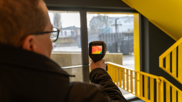 Na zdjęciu widać tyłem osobę, która trzyma w ręku przyrządu do badania termowizyjnego i wykonuje pomiary.