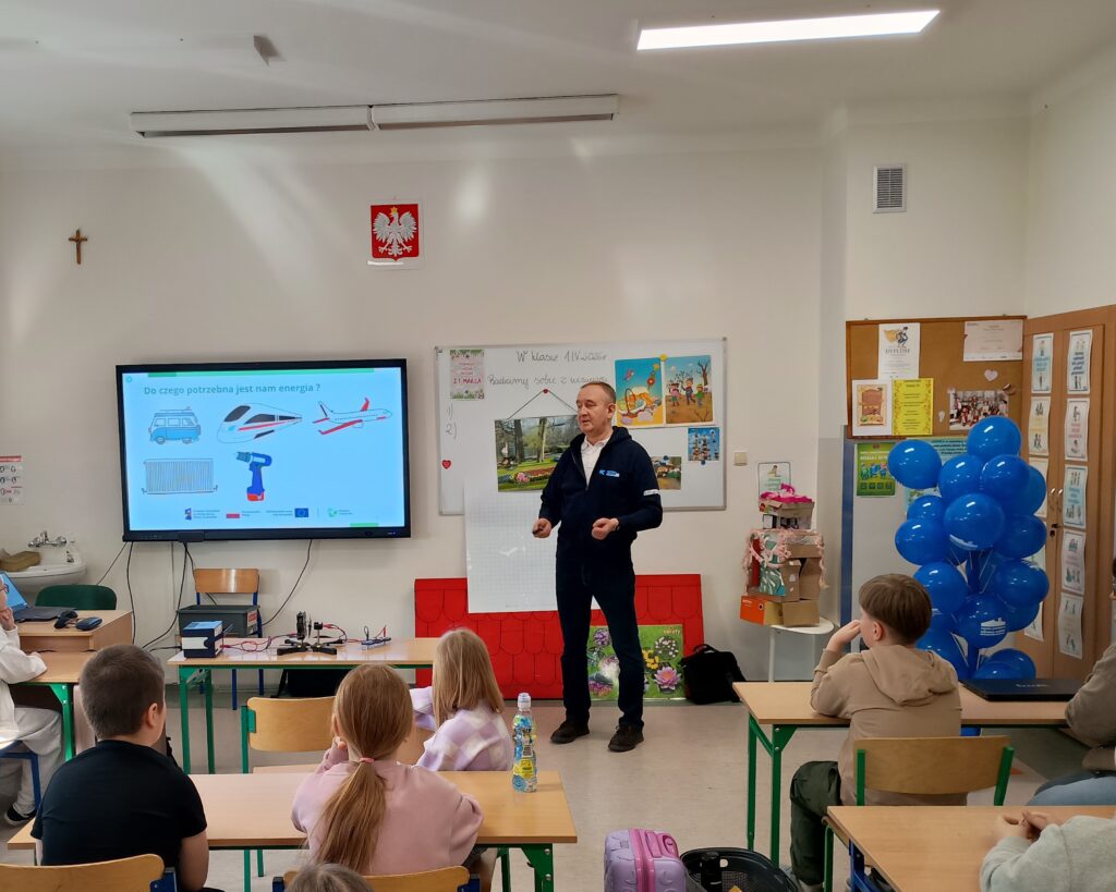 Na zdjęciu widzimy zajęcia w szkolnej klasie. Osoba prowadząca zajęcia stoi naśrodku sali i omawia prezentację, która wyświetlona jest na dużym telewizorze.