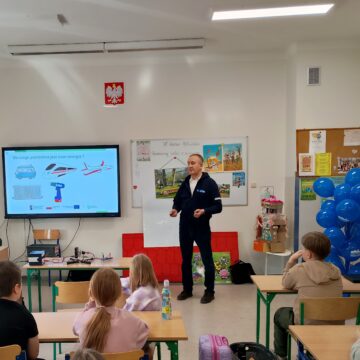 Na zdjęciu widzimy zajęcia w szkolnej klasie. Osoba prowadząca zajęcia stoi naśrodku sali i omawia prezentację, która wyświetlona jest na dużym telewizorze.