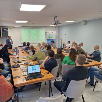 Na zdjęciu widzimy salę, na której przy długich stołach siedzą osoby i słuchają prelekcji wygłaszanej przez prowadzącego, który wyświetla na ekranie prezentację.